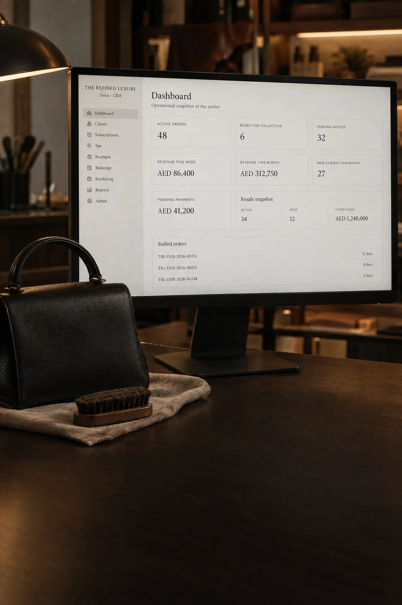 A monitor in a leather-care atelier showing The Refined Luxury operations dashboard, beside a black leather handbag