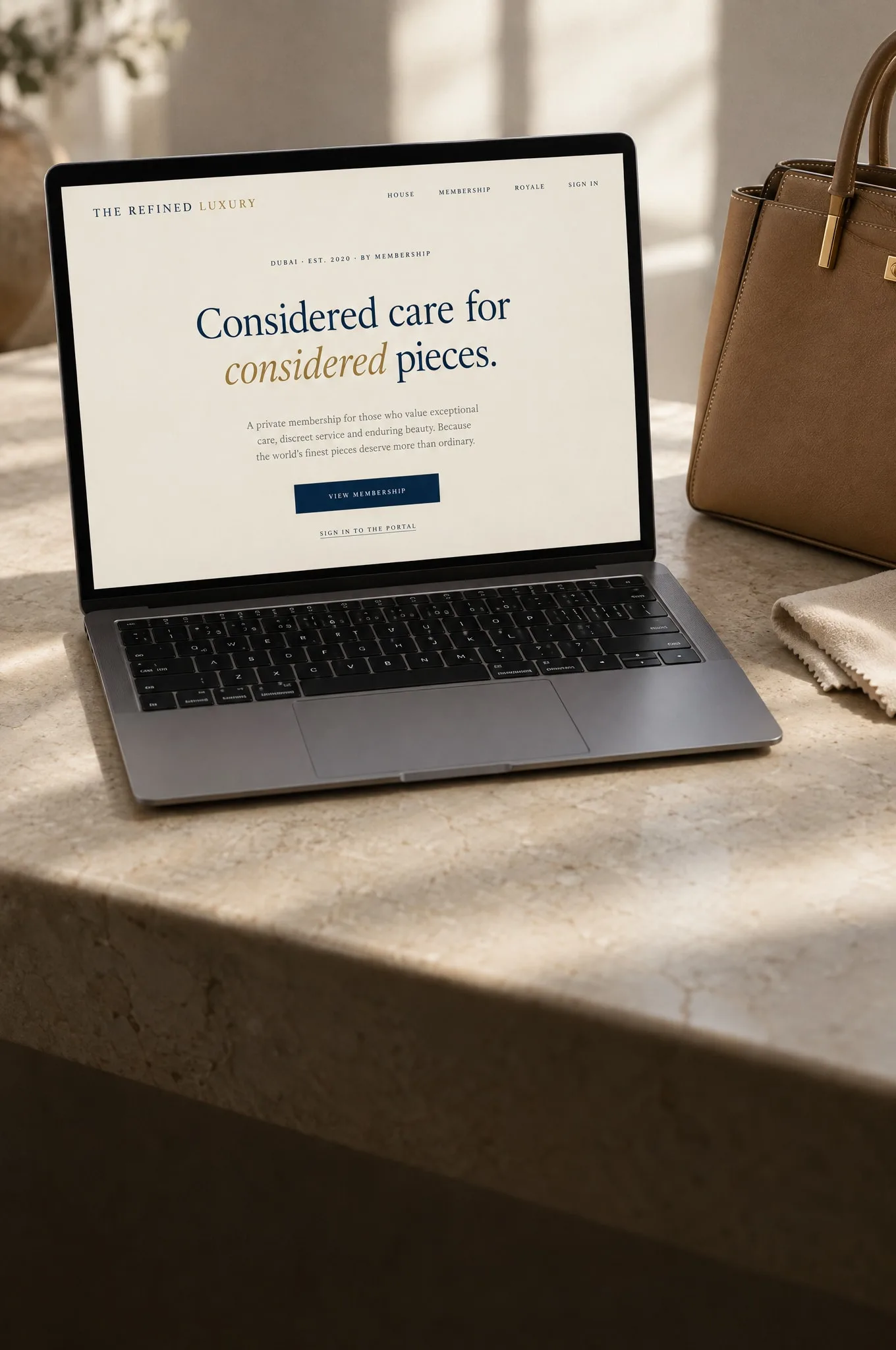 A laptop on a stone surface showing The Refined Luxury membership site, beside a tan leather handbag