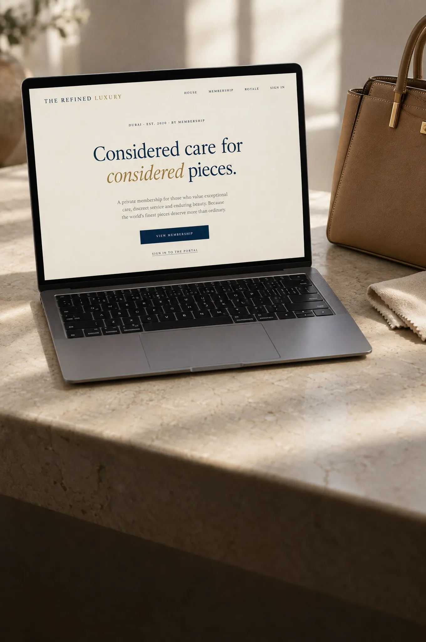 A laptop on a stone surface showing The Refined Luxury membership site, beside a tan leather handbag